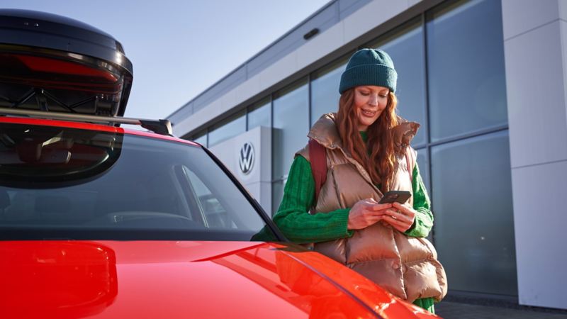 Eine Frau schaut auf ihr Smartphone und steht an der Fahrertür eines roten VW mit schwarzer Dachbox.