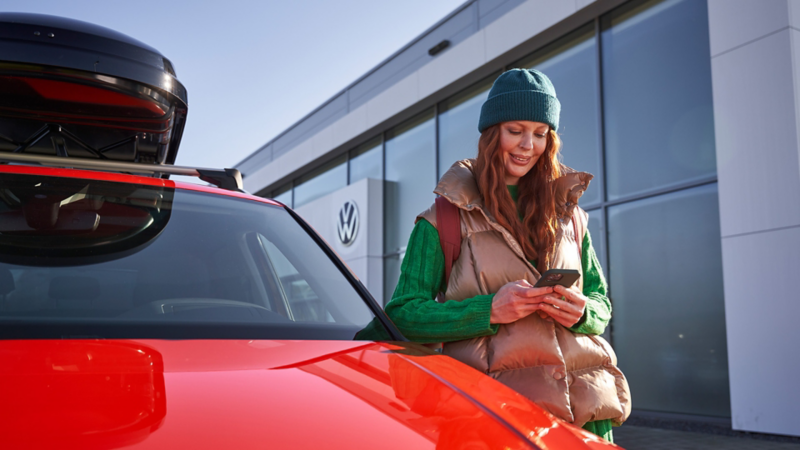 Eine Frau schaut auf ihr Smartphone und steht an der Fahrertür eines roten VW mit schwarzer Dachbox.