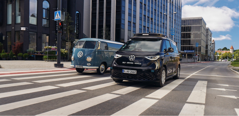 Ein selbstfahrender VW ID. Buzz und ein VW T1 Bulli fahren nebeneinander durch eine Stadt.