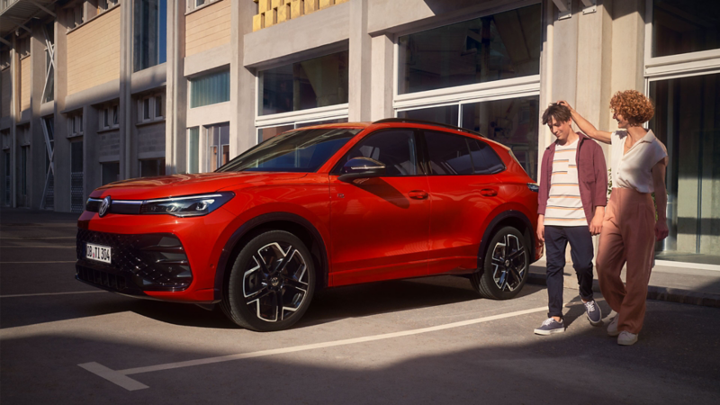 Eine Frau und ein Mann verlassen ein Gebäude mit großen Glasfenstern und gehen an einem roten VW Tiguan vorbei.