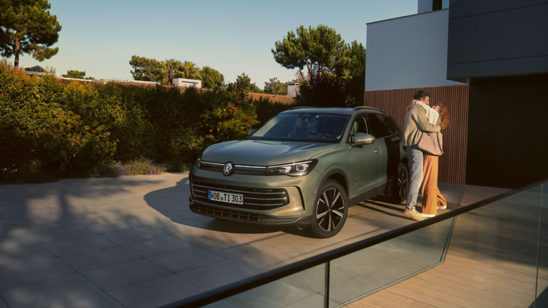 Vue de face d'un VW Tiguan vert garé dans l'allée d'un immeuble résidentiel moderne. Un jeune couple se tient à proximité et s’embrasse.