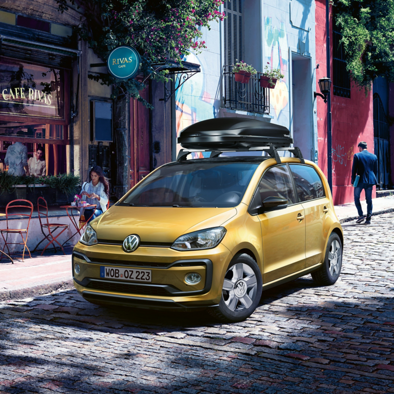 Ein goldgelber VW up! mit Dachbox parkt vor einem Straßencafé.