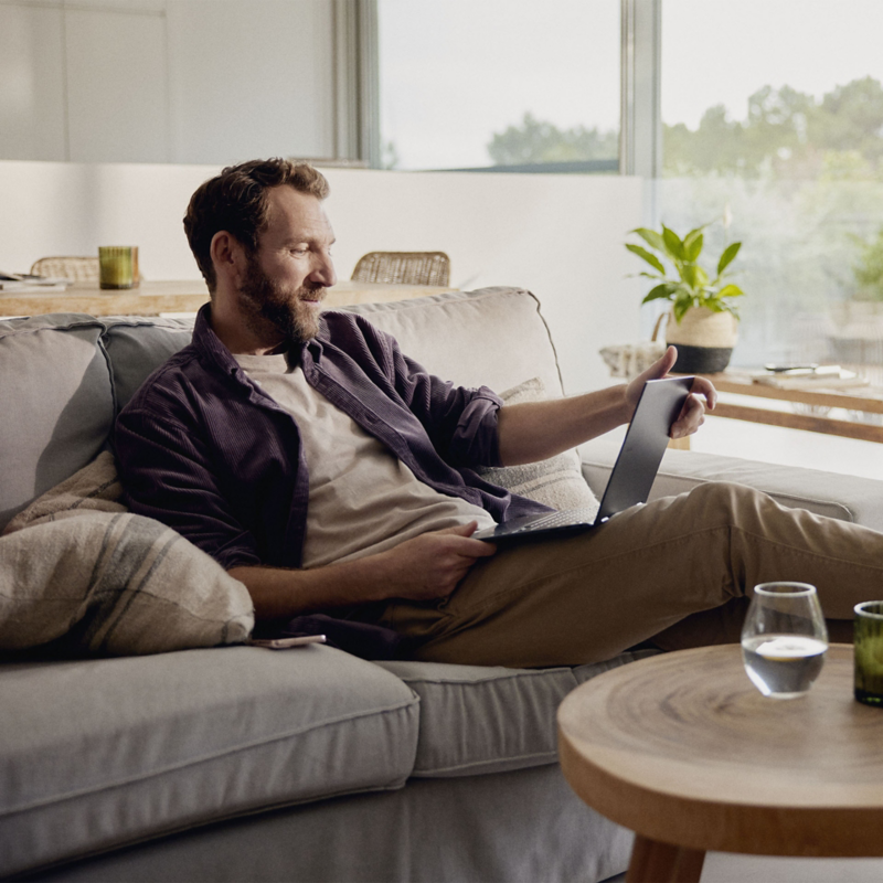 Ein Mann sitzt gemütlich auf dem Sofa, hat die Füße hochgelegt und blickt auf seinen Laptop.