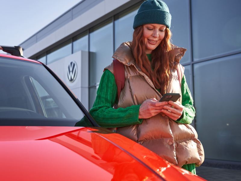Eine Frau schaut auf ihr Smartphone und steht an der Fahrertür eines roten VW mit schwarzer Dachbox.