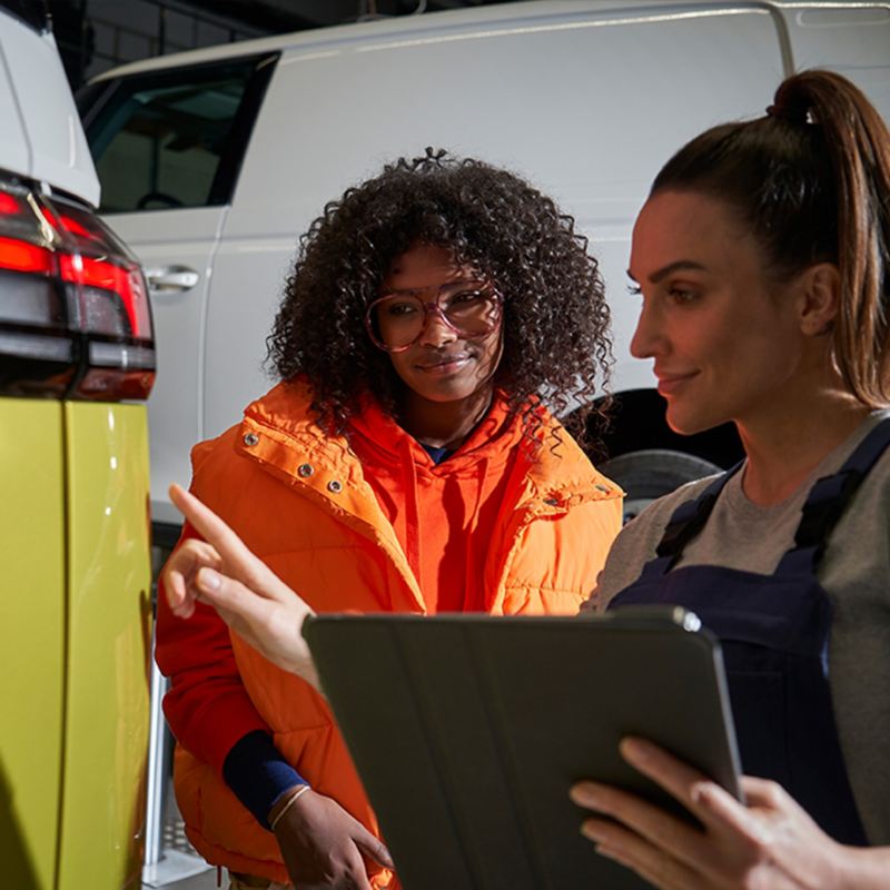 Eine Mechanikerin steht mit einer Frau und einem Tablett-PC am Heck eines VW ID. Buzz und erklärt etwas.