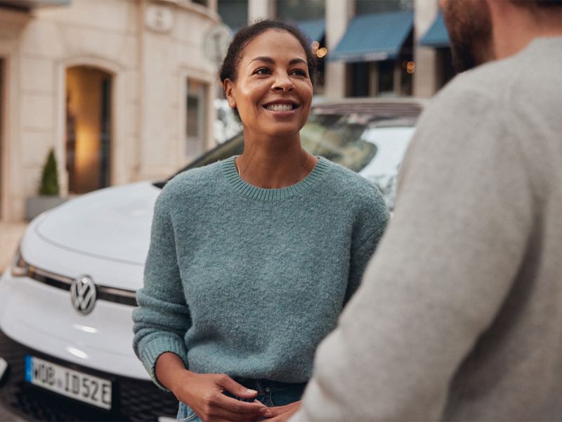 Eine Frau steht vor einem weißen VW und unterhält sich mit einem Mann.