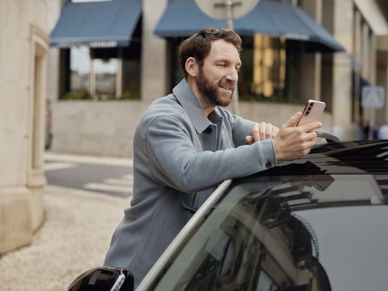 Ein Mann lehnt an der Beifahrerseite eines Volkswagen und schaut auf sein Smartphone.