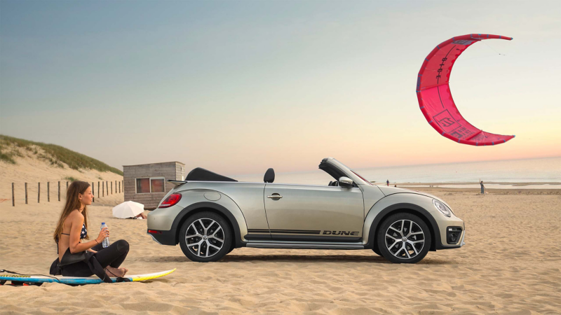 VW Beetle convertible on beach with woman sat next to it