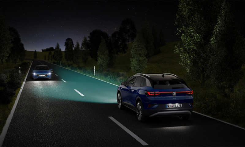 A blue VW ID.4 drives down a street with its lights on at night.