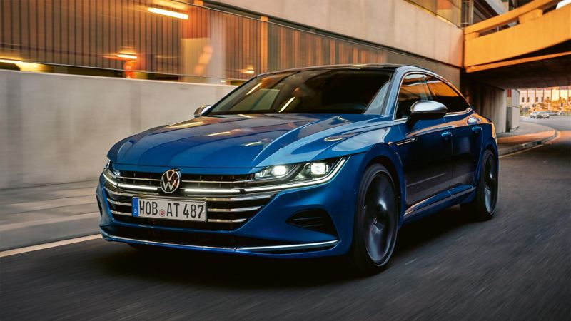 A blue Volkswagen Arteon sedan drives through a city at sunset