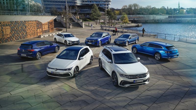 Sju vita och blåa Volkswagenmodeller uppställda i cirkel på parkering vid vatten
