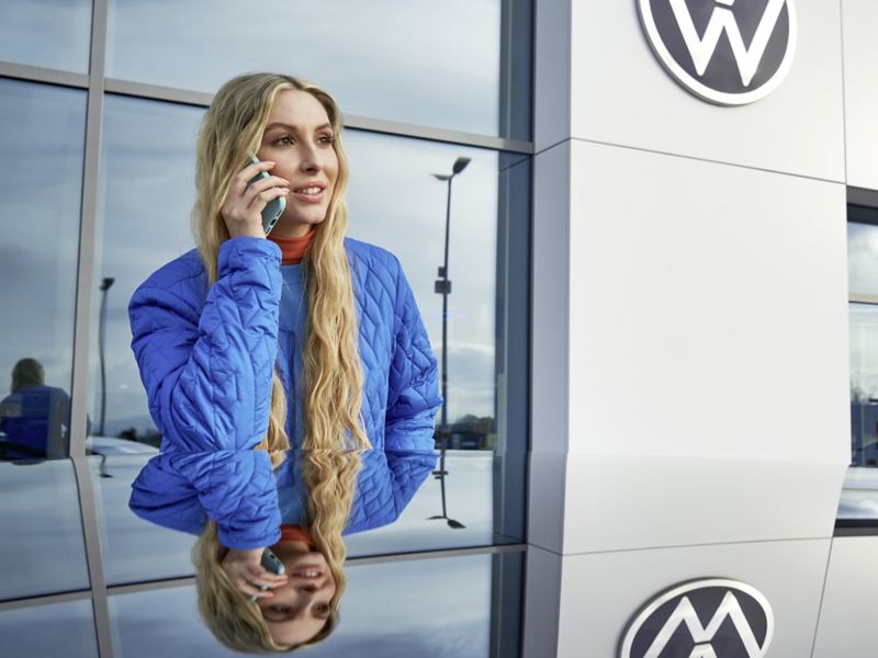 Eine Frau telefoniert und steht vor einem Gebäude mit Volkswagen Logo.