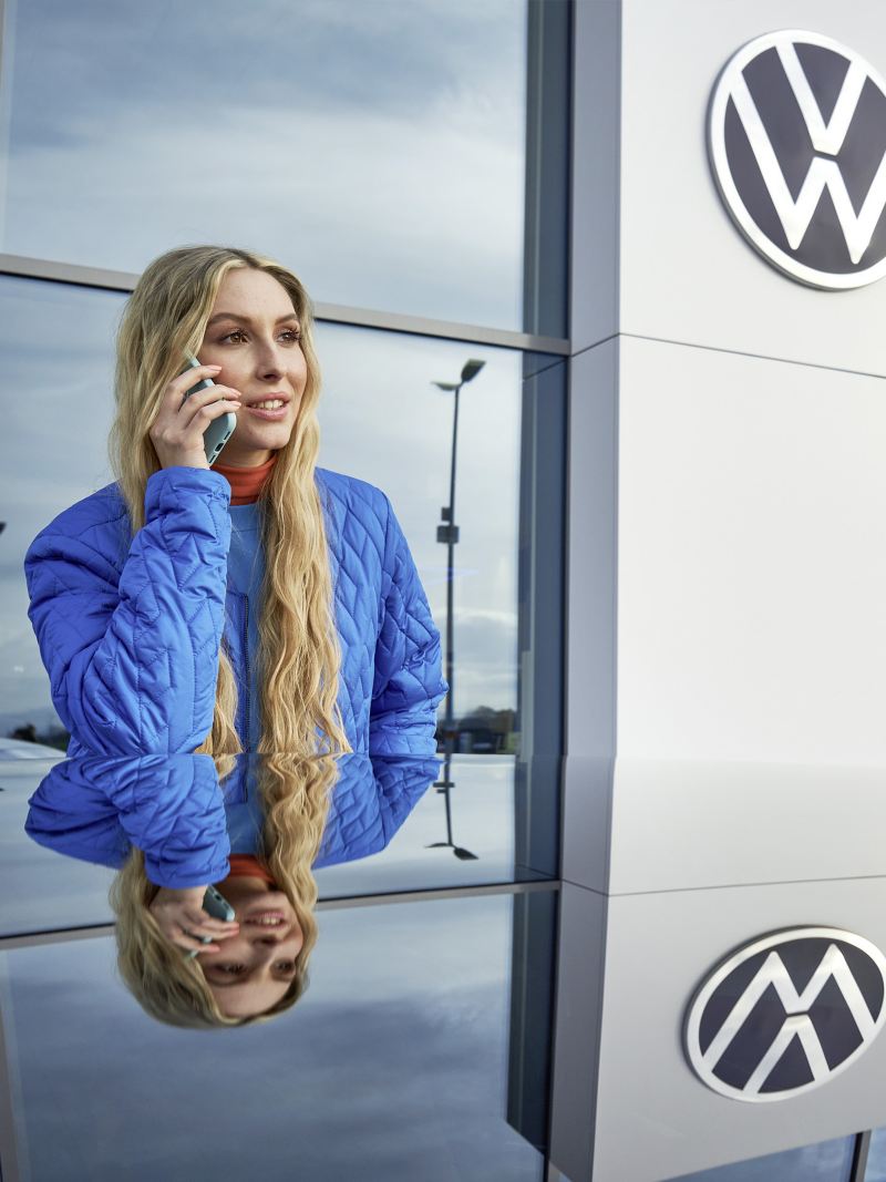 Eine Frau telefoniert und steht vor einem Gebäude mit Volkswagen Logo.