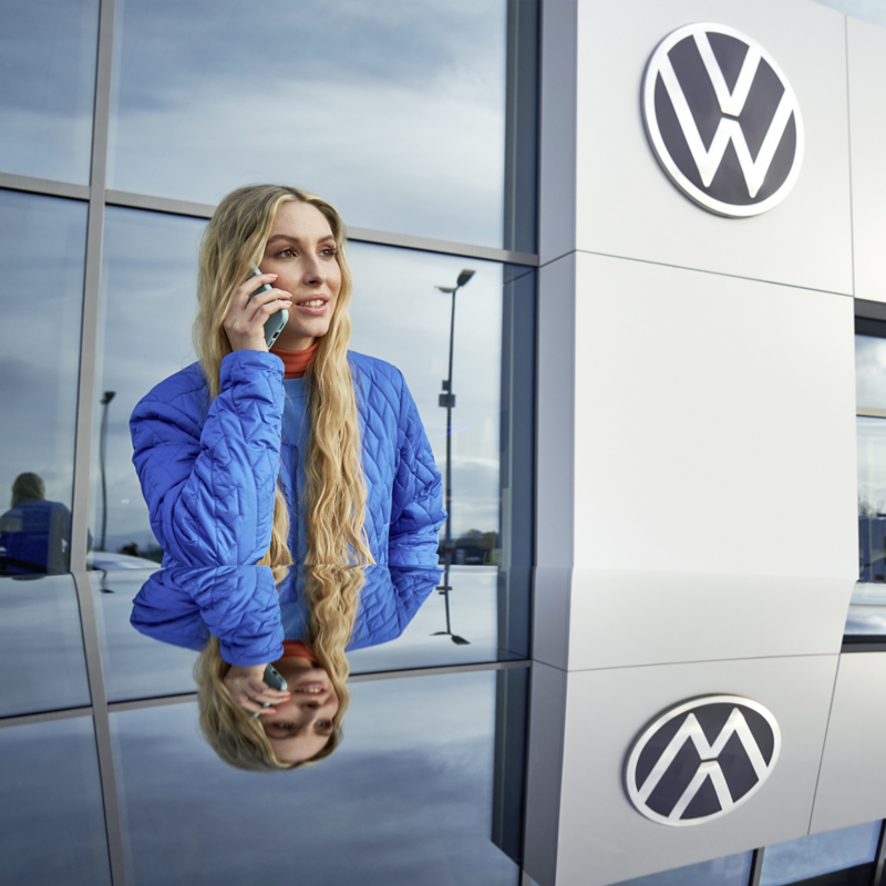 Eine Frau telefoniert und steht vor einem Gebäude mit Volkswagen Logo.