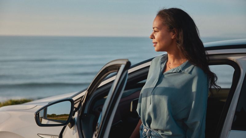 Eine Frau steht an der offenen Fahrertür eines VW, der am Meer geparkt ist und blickt in die Sonne.