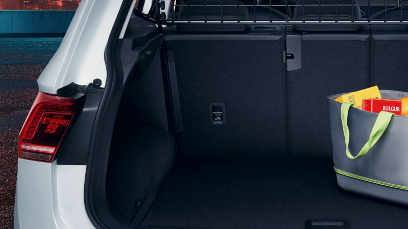 Le coffre ouvert d'une Volkswagen Tiguan, montrant l'espace de chargement vide et une sacoche de courses grise avec des paquets colorés à l'intérieur. Le feu arrière gauche de la voiture est visible.