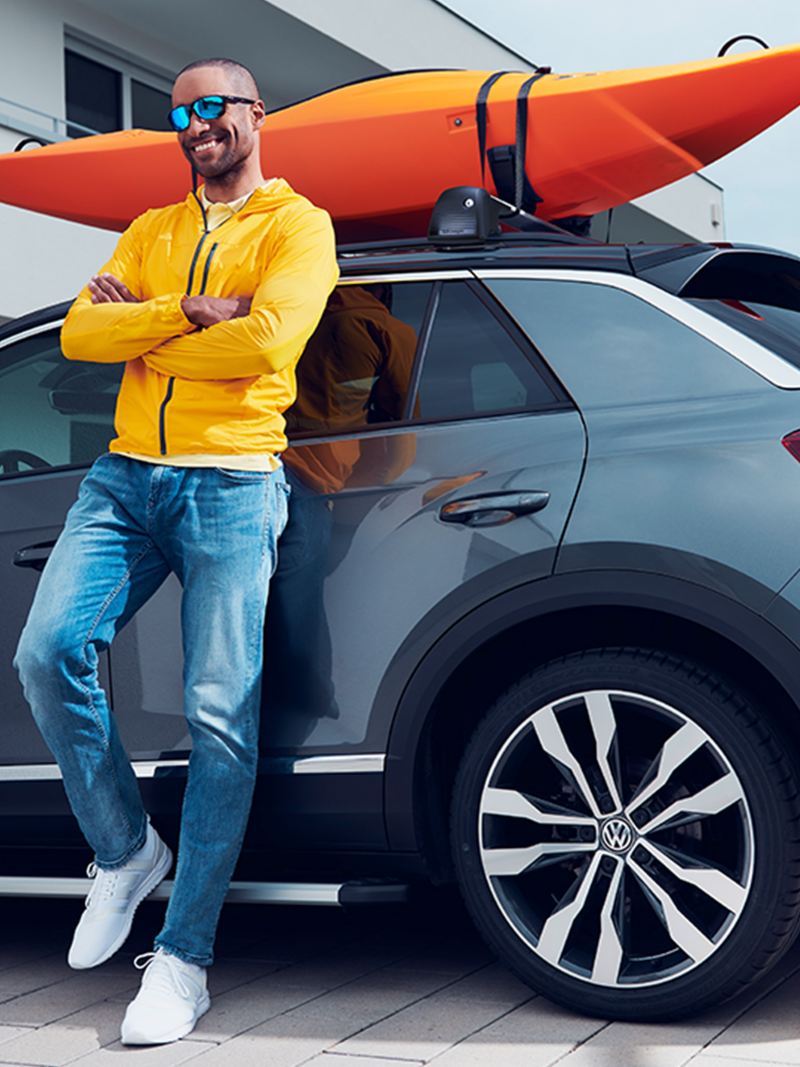 Ein Mann lehnt an grauem VW mit orangefarben Kajak auf dem Dachgepäckträger.