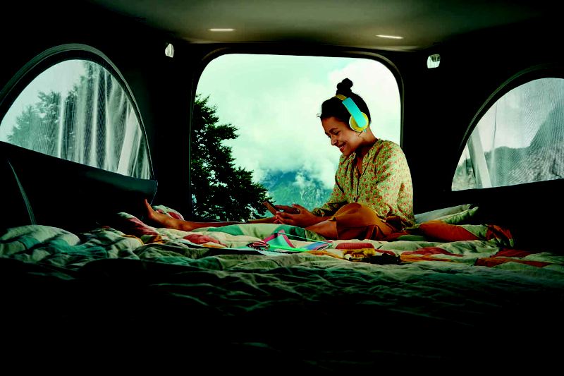 外國女生坐在California車內面帶微笑的正在滑手機，車內鋪著棉被看起來相當舒適