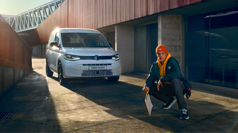 Biały Volkswagen Caddy Cargo zaparkowany przy nowoczesnym budynku oraz kucający obok mężczyzna w pomarańczowej czapce i żółtej bluzie z podkładką z dokumentami.