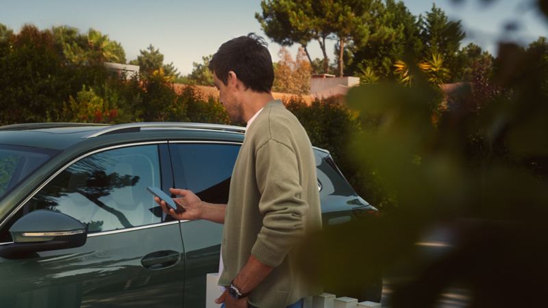Ein junger Mann auf der Fahrerseite des dunkelgrünen VW Tiguan bedient den optionalen Park Assist Plus über sein Smartphone.