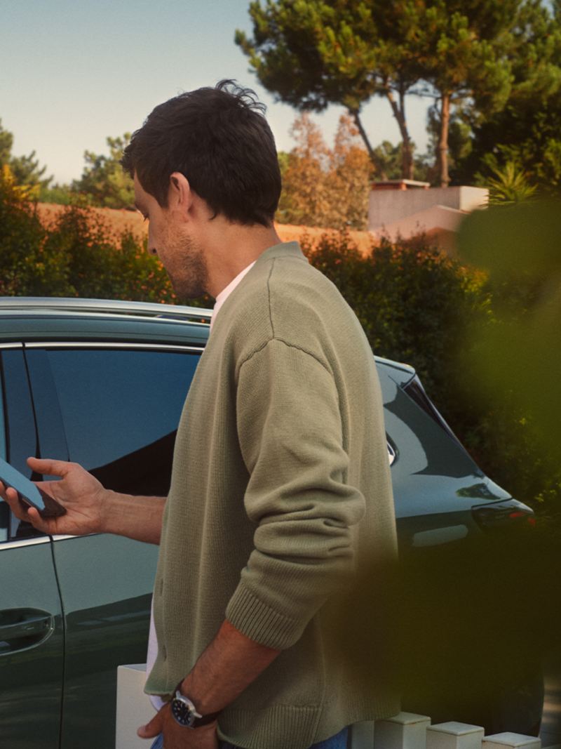 A young man on the driver side of the dark green VW Tiguan operates the optional Park Assist Plus from his smartphone.