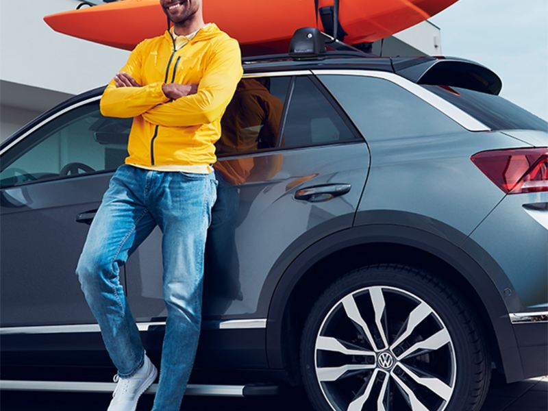 Ein Mann lehnt an grauem VW mit orangefarben Kajak auf dem Dachgepäckträger.