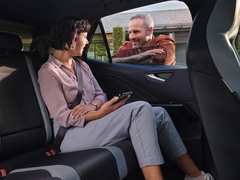 Eine Frau sitzt auf der Rückbank eines VW. Sie unterhält sich durch das offene Fenster mit einem Mann, der von außen angelehnt steht.