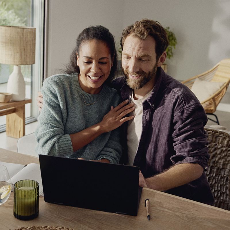 Ein Paar sitzt Zuhause am Tisch und schaut lächelnd gemeinsam auf einen Laptop-Bildschirm.