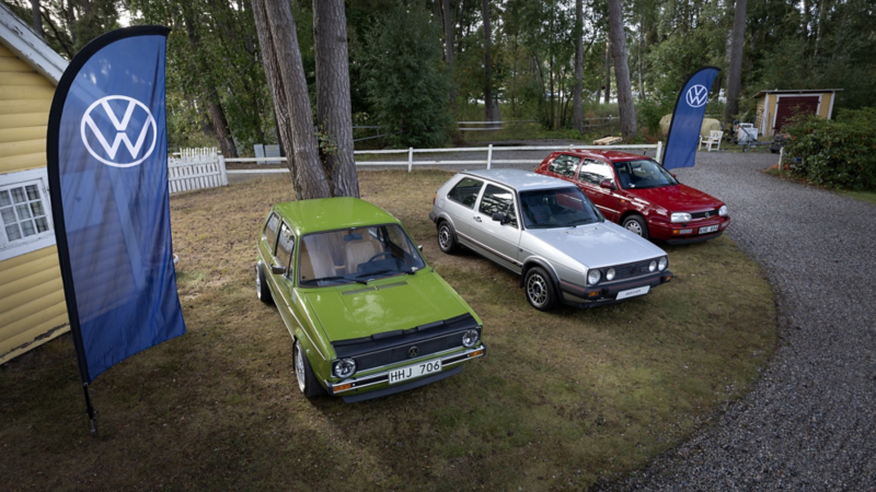 Tre klassiska Volkswagen Golf står parkerade på gräs i en parkmiljö, med Volkswagen-flaggor och träd i bakgrunden.