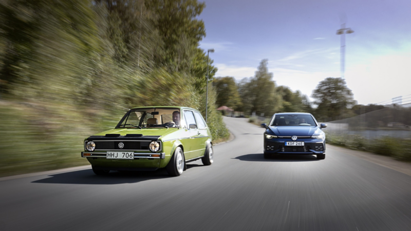 En klassisk grön Volkswagen Golf och en modern blå Volkswagen Golf kör sida vid sida på en landsväg i grön omgivning.