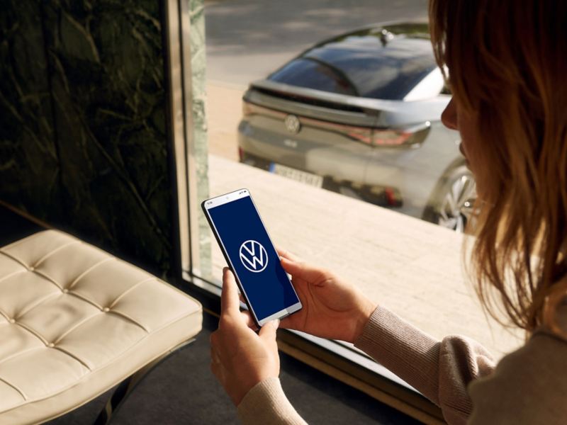 Eine Frau sitzt hinter einem Fenster und blickt auf ihr Smartphone, auf dem ein VW Logo und hinter dem Fenster im Hintergrund ein VW zu sehen ist. 