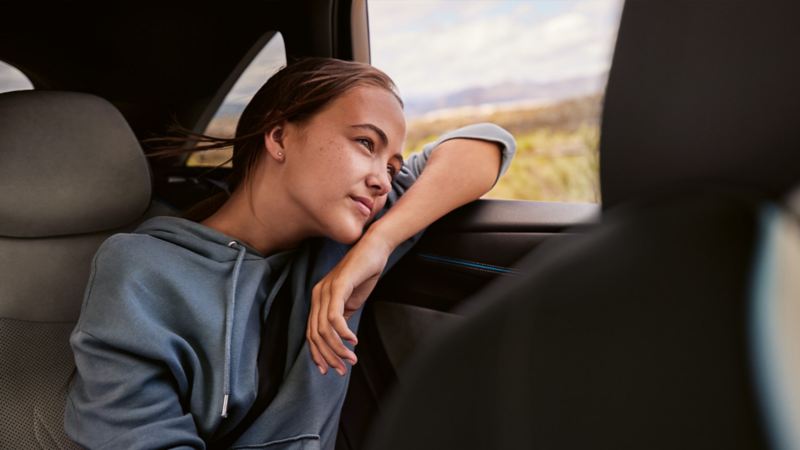 Ein junges Mädchen sitzt auf der Rückbank eines VW und blickt durch das offene Fenster.