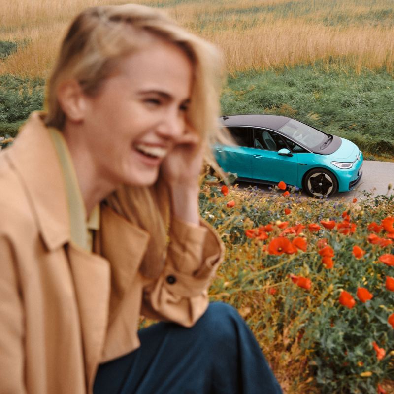 Una mujer con una elegante gabardina se ríe, al fondo se ve un VW ID.3
