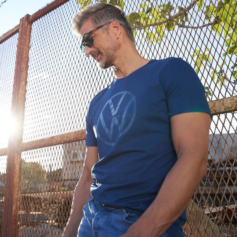 Ein Mann in einem T-Shirt aus der Volkswagen Kollektion lehnt an einem hohen Zaun