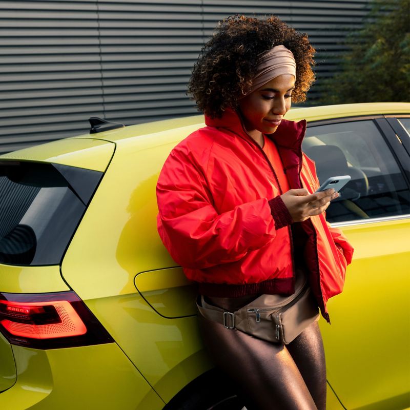 Eine Frau lehnt an ihrem VW Auto und bucht einen Driving Experience Termin