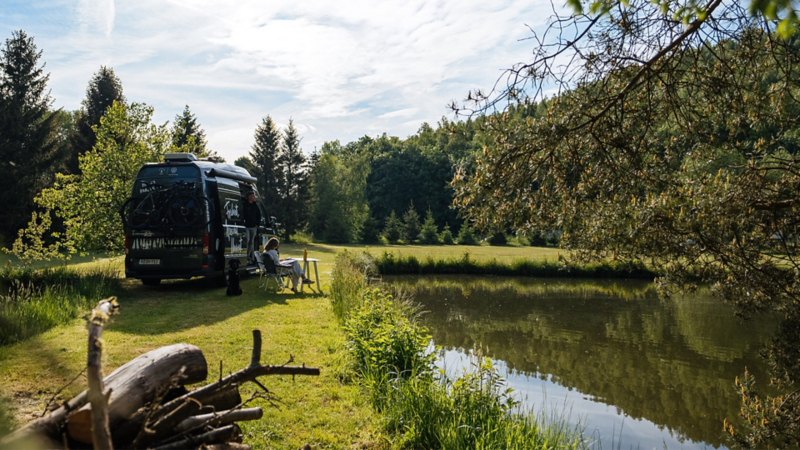 Un camper Grand California est garé sur une prairie verte au bord d’un lac. Une personne est assise à côté, détendue, sur une chaise de camping.