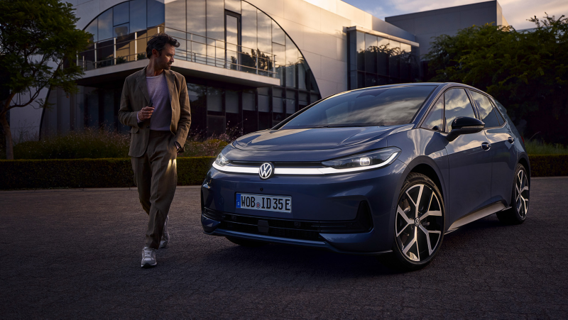Blauer VW ID.3 Neo parkt vor modernem Glasgebäude, eine gehende Person daneben, Straße bei Abendlicht.