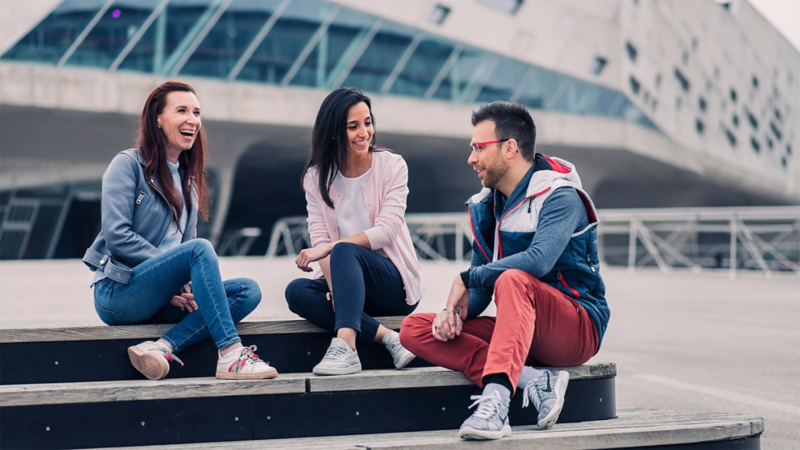 Two young women and a young man sitting on steps next to a VW building