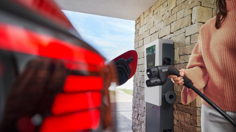 Eine Frau steckt den Ladestecker in ein rotes VW Elektrofahrzeug an einer DC-Wallbox.