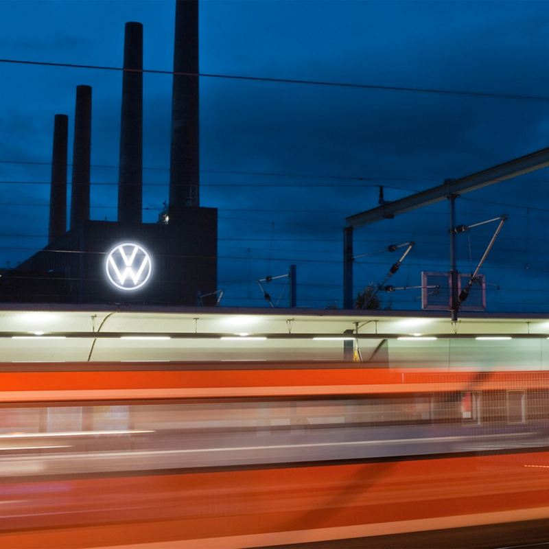 Ein fahrender Zug und im Hintergrund das Volkswagen Werk Wolfsburg bei Nacht