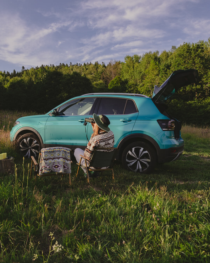 Turkusowy Volkswagen na łące przy lesie, obok na krześle siedzi kobieta