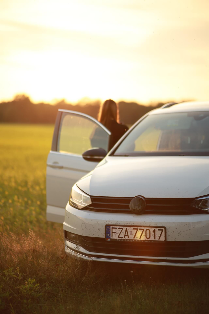 Biały Volkswagen na łące przy zachodzie, obok stoi kobieta
