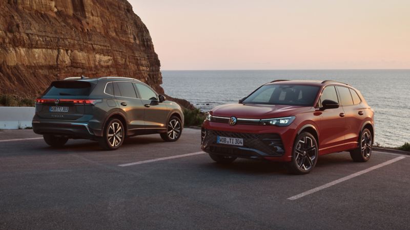 Two VW Tiguan in dark green and red are parked in front of a rocky coastal landscape.