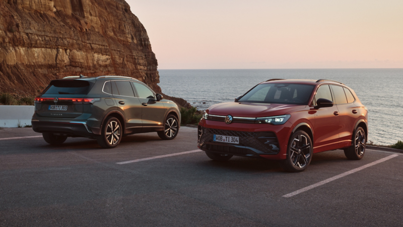 Two VW Tiguan in dark green and red are parked in front of a rocky coastal landscape.