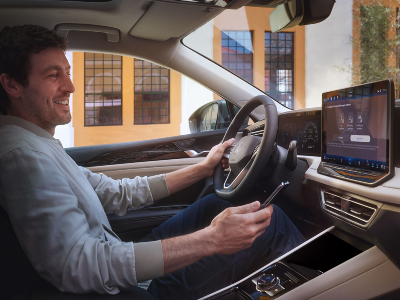 Ein lächelnder Mann sitzt, mit Smartphone in der Hand, im VW Tiguan und schaut auf das Infotainment-System.