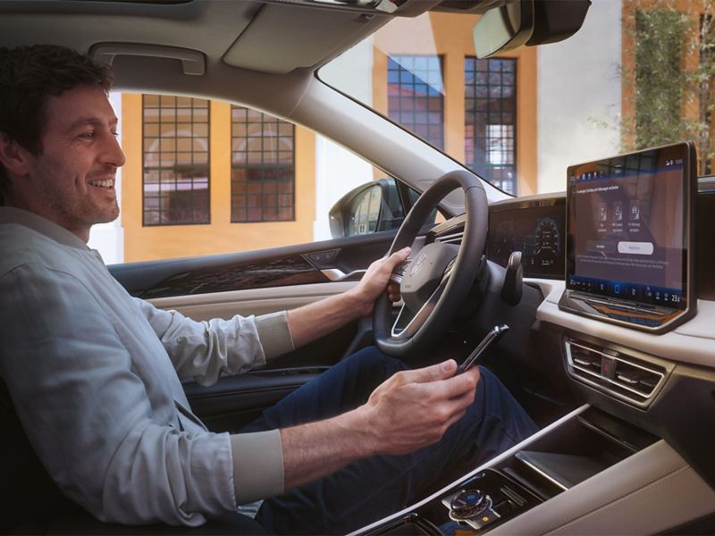 Kabinen i en VW Tiguan. En mand på førersædet styrer VW Connect Plus-appen (ekstraudstyr) med sin smartphone.