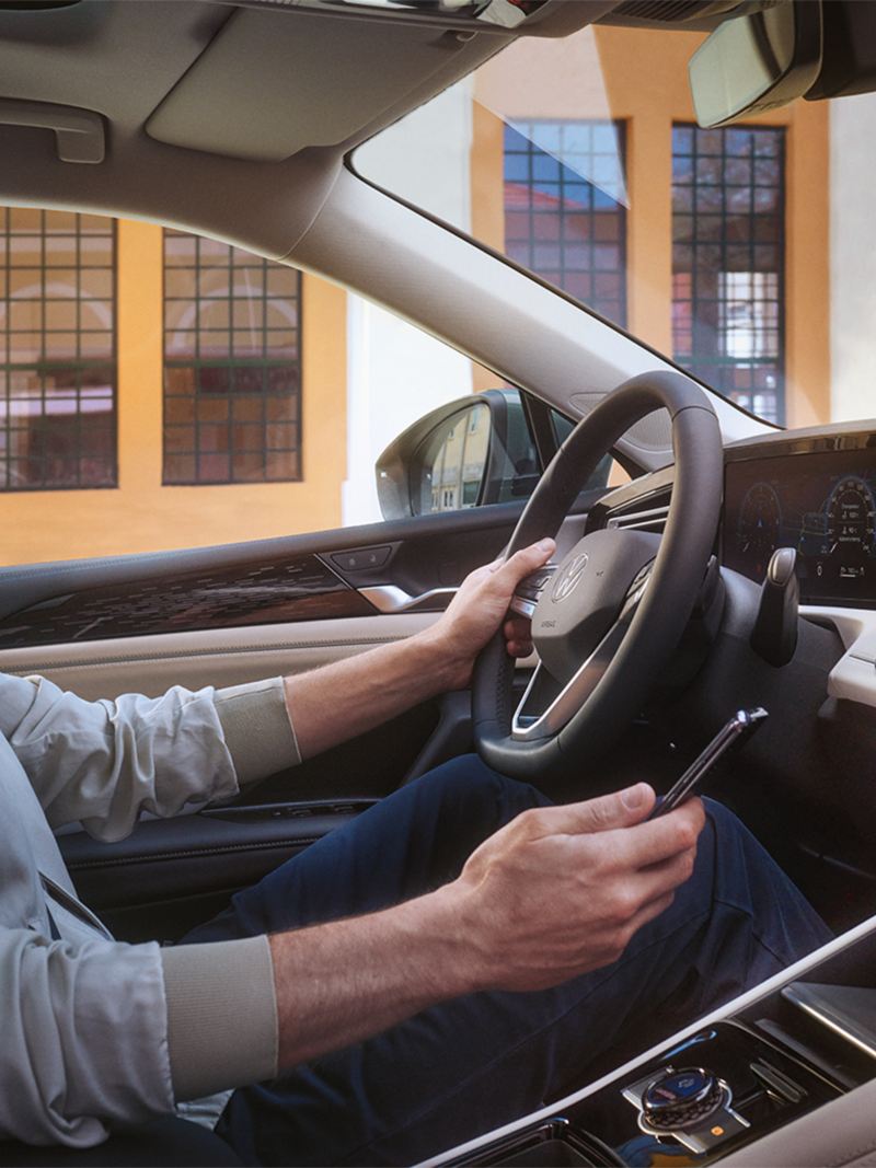 Vue de l’intérieur d’un VW Tiguan. Un homme assis sur le siège conducteur utilise son smartphone pour contrôler l’application We Connect Plus disponible en option.