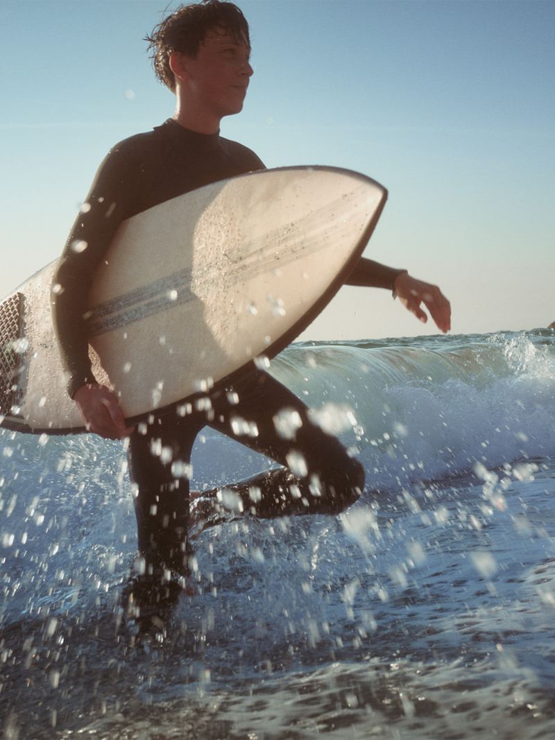 Plaża, młody mężczyzna z deską surfingową brodzi po sięgającej do kolan wodzie.