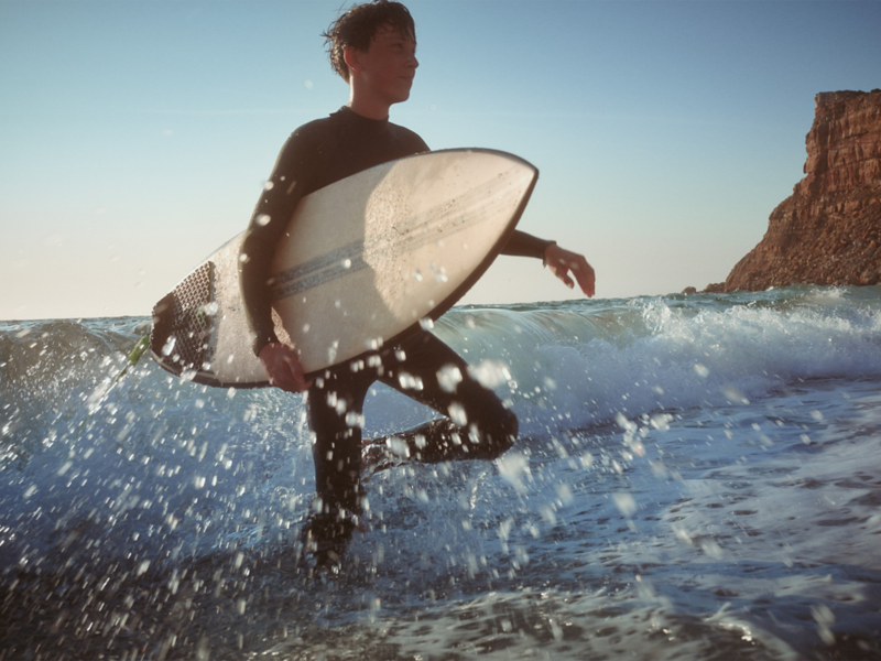 Plaża, młody mężczyzna z deską surfingową brodzi po sięgającej do kolan wodzie.