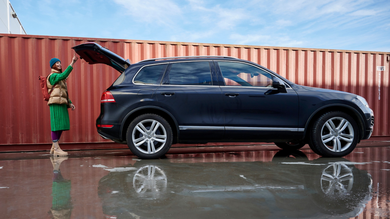 Woman stands at the luggage compartment of a black VW Touareg 2 and presses the button to automatically close the tailgate